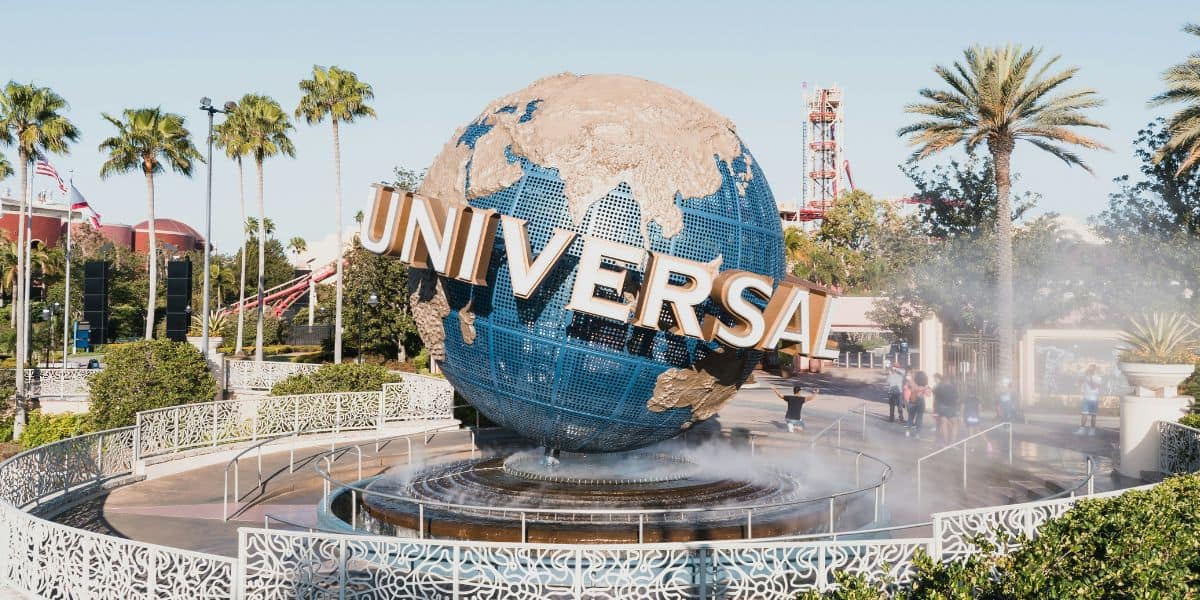 A large rotating globe with the word "Universal" prominently displayed stands as the iconic entrance landmark for Universal Land. It is surrounded by lush greenery, mist, and white fences, with palm trees and park attractions visible in the background.