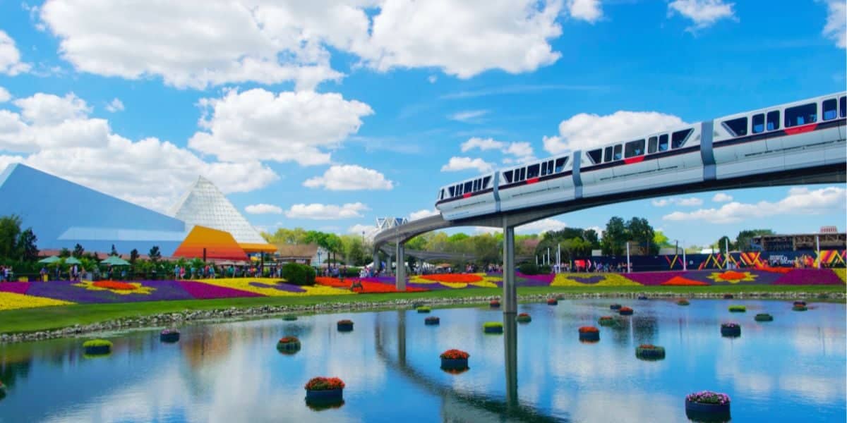 A monorail travels above a beautiful, landscaped garden with vibrant flowers arranged around a serene pond. In the background are modern, geometric buildings beneath a bright blue sky with scattered white clouds. Despite the bustling Walt Disney World crowds, people stroll and enjoy the sunny day.