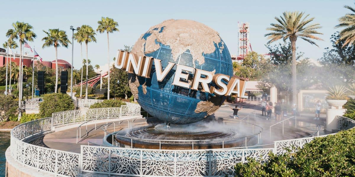 The Universal globe with gold letters stands prominently at the entrance of Universal Orlando Resort. Surrounding the fountain are palm trees, lush greenery, and mist from the water. In the background, theme park attractions and animated animals are visible.