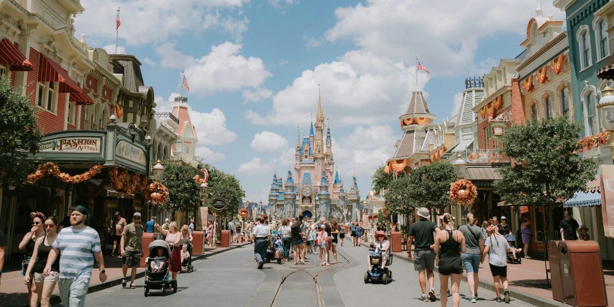 A vibrant street scene at Disney theme park features people strolling along a main avenue adorned with festive fall decorations. Shops line both sides, and a castle looms majestically in the background under a bright, cloudy sky. Nearby, the entrance to Peter Pan’s Flight draws excited visitors.