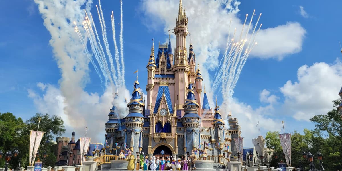 A large, magical castle with blue and gold accents stands majestically under a bright blue sky with scattered clouds. Surrounding the castle are fireworks, and at the base, a group of costumed characters engage with onlookers.