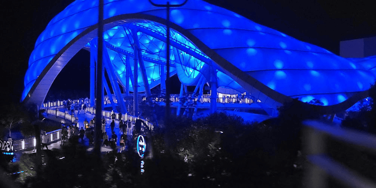 Nighttime view of a futuristic, blue-lit pedestrian bridge bustling with people, featuring a distinctive arched design and glowing light spheres.