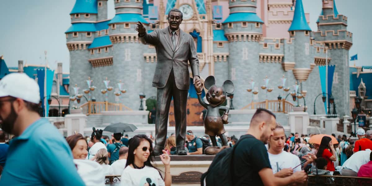 A crowd of people gather around the statue of Walt Disney and Mickey Mouse in front of Cinderella's Castle at Disney World. The castle is adorned in blue and gold spires, while the people wear various casual outfits, enjoying the lively atmosphere of the park.