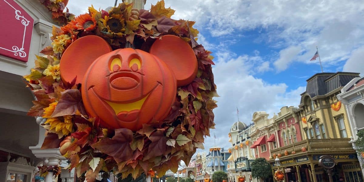 Festive Halloween decorations at Disney World showcase a large Mickey Mouse pumpkin surrounded by autumn leaves. Colorful buildings line the street under a partly cloudy sky, with an American flag visible in the background, capturing the vibrant spirit of the season.