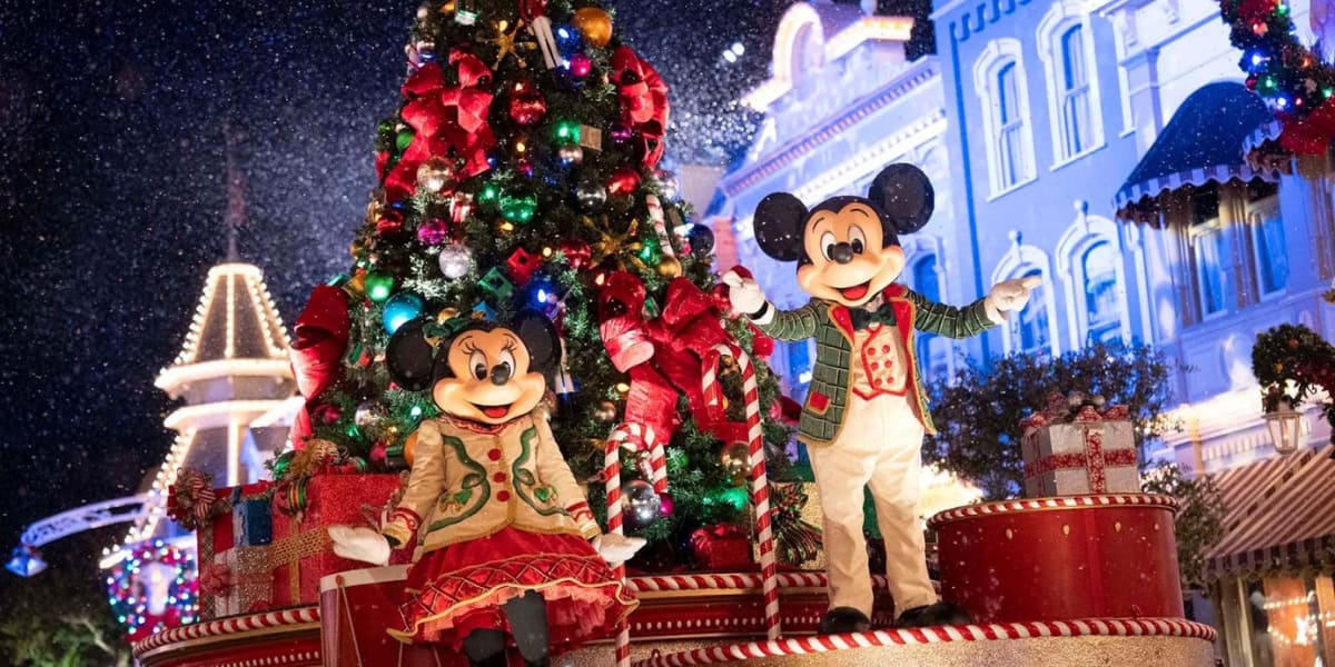 Mickey and Minnie Mouse, dressed in festive holiday attire, stand in front of a large, decorated Christmas tree. They are surrounded by colorful, wrapped gifts, and twinkling lights adorn the scene. Snow appears to be gently falling in the nighttime setting.