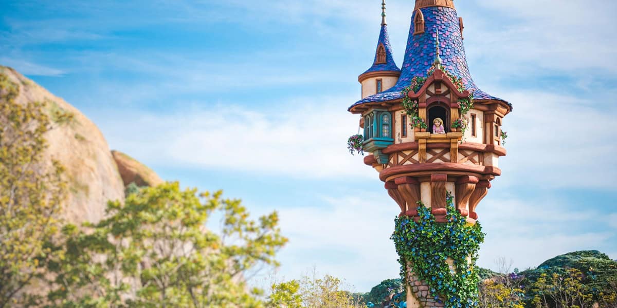 A whimsical, colorful tower stands tall amidst a backdrop of trees and rocks. The tower has a conical roof, intricate details, and ivy climbing its base. A figure stands in a window near the top, looking out into the distance, against a clear blue sky.