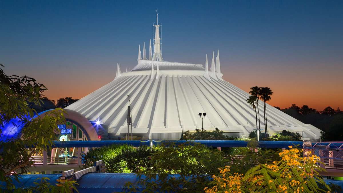 The image features Space Mountain, a distinctive white futuristic building with spire-like structures at Disney World, at dusk with a colorful sunset sky and foreground lush greenery.