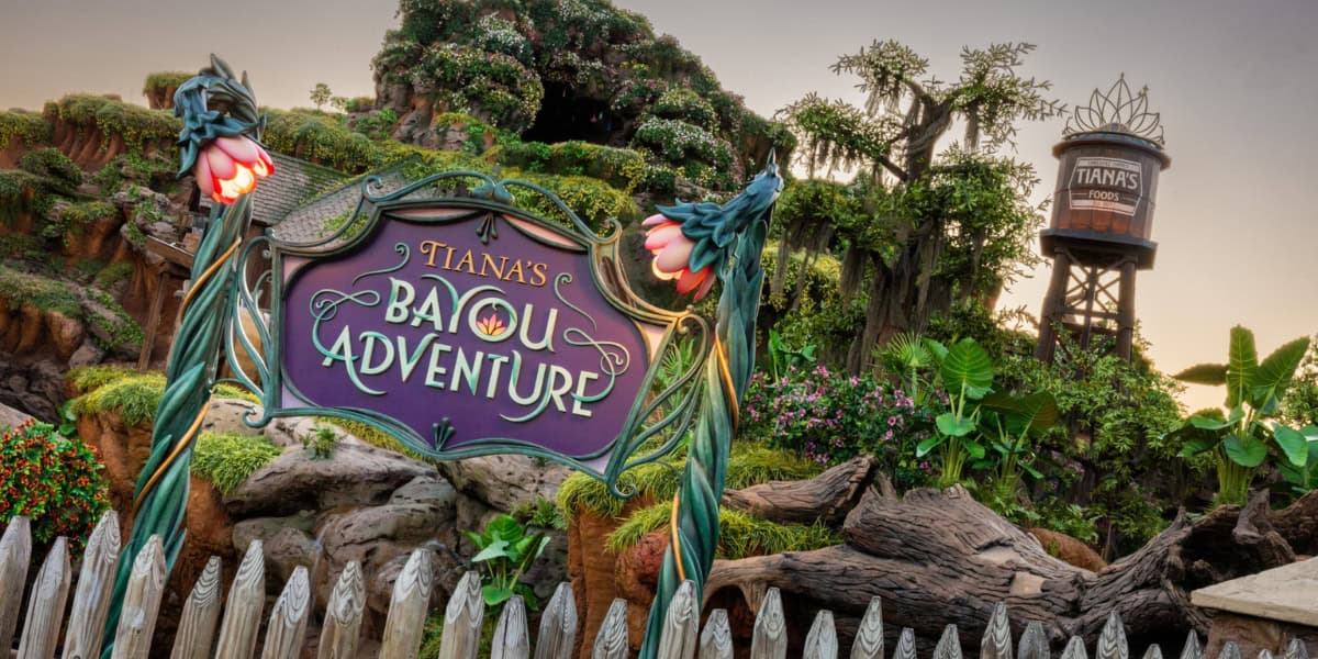 The image shows the entrance sign for "Tiana's Bayou Adventure" with a water tower in the background that also reads "Tiana's Foods."