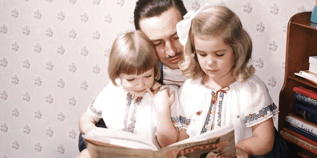 A man with a mustache reads a book to two young children, both girls, sitting on his lap. The girls are dressed in matching white outfits with colorful embroidered patterns. They are indoors, against a wallpapered backdrop.