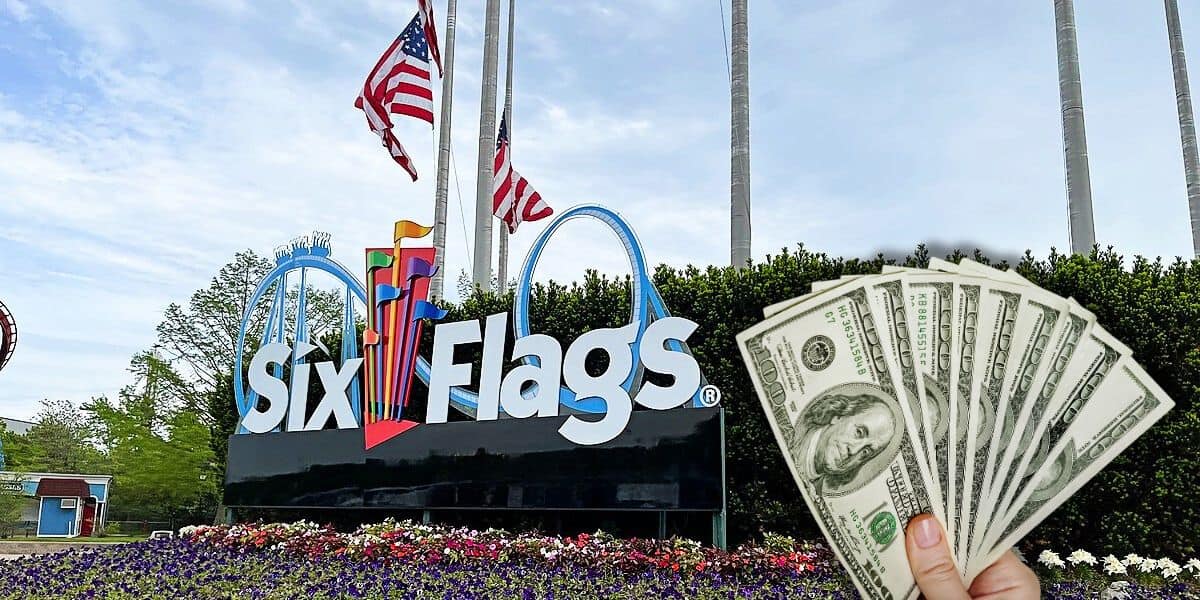 A person holds a fan of U.S. dollar bills in front of a Six Flags amusement park entrance sign, reminiscent of a Cedar Fair park. The sign features a colorful logo with flags, and American flags are flying on flagpoles behind it. Surrounding the sign are manicured shrubs and flower beds.