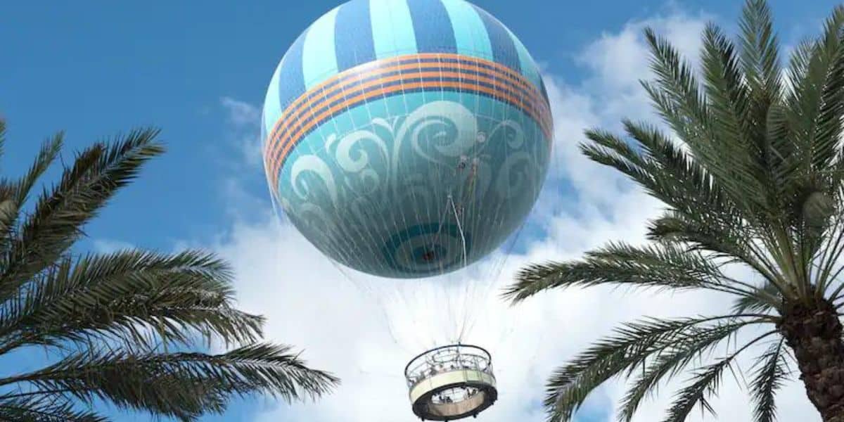 A large, colorful hot air balloon with swirling patterns floats against a blue sky, reminiscent of a whimsical Disney World scene, framed by palm trees.