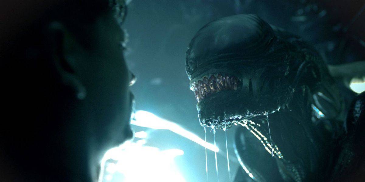 A close-up of an alien creature with sharp teeth and a shiny, textured head, facing a person in a dark, Disney+ atmospheric setting. A bright light glows in the background, highlighting the creature's menacing expression.