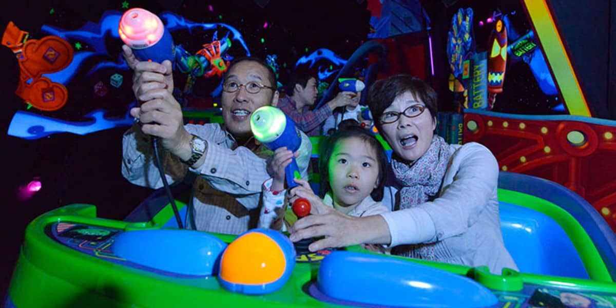 A family enjoys an interactive ride, with a child and two adults shooting light guns in a dimly lit, colorful arcade-style environment. they appear excited and engaged in the fun activity.