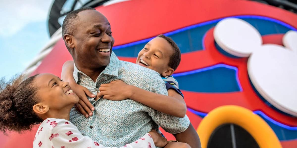 A joyful man embraces two smiling children, one girl and one boy, in front of a vibrantly colored structure featuring the iconic Mickey Mouse logo on the Disney Cruise Line