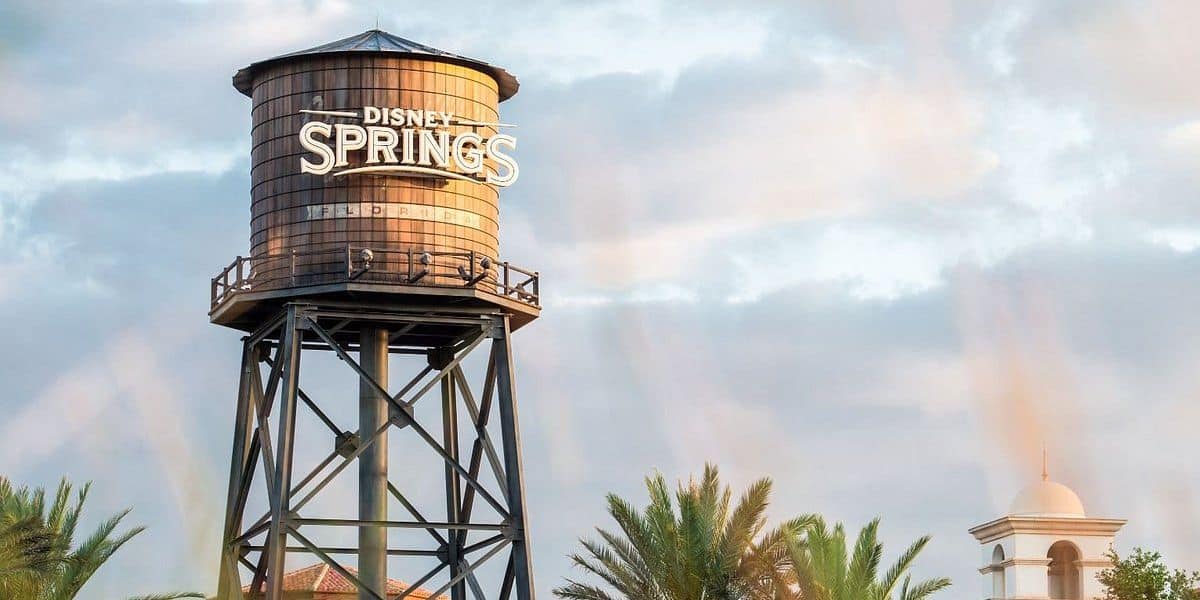 An image of the Disney Springs water tower on a cloudy day, with light rays peaking through the clouds, and a clear sky in the background featuring an advertisement for Chicken Guy sauces.