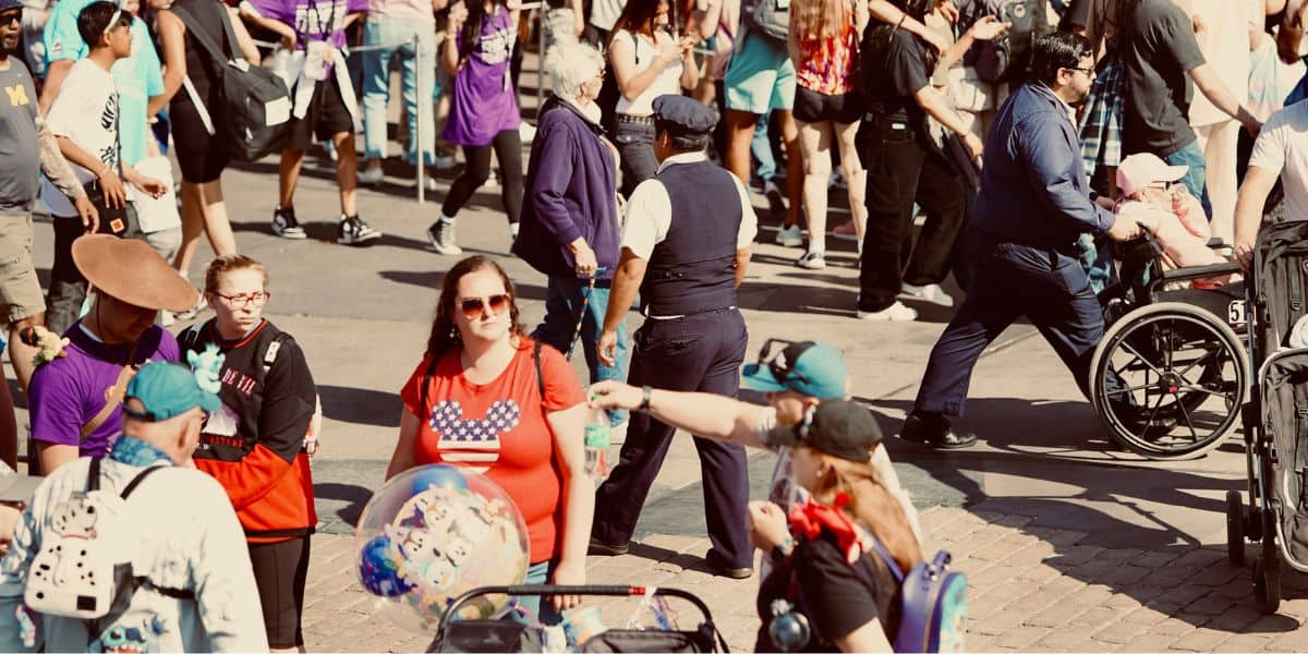 A busy street scene with a diverse group of people walking and interacting. A police officer stands in the middle, monitoring the crowd. Some people are wearing backpacks, hats, and pushing strollers, including a person in a wheelchair and a child in another stroller amidst whispers of a Disney evacuation.