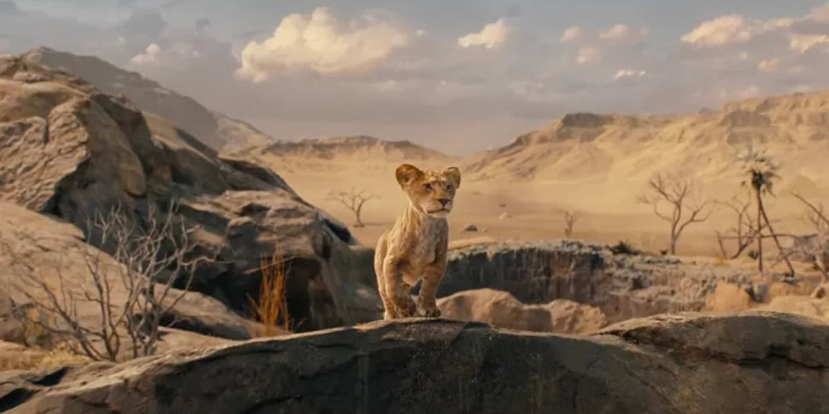 A young lion cub stands atop a rocky ledge overlooking a vast, arid savannah landscape with sparse vegetation and distant mountains under a clear sky, in The Lion King: Mufasa