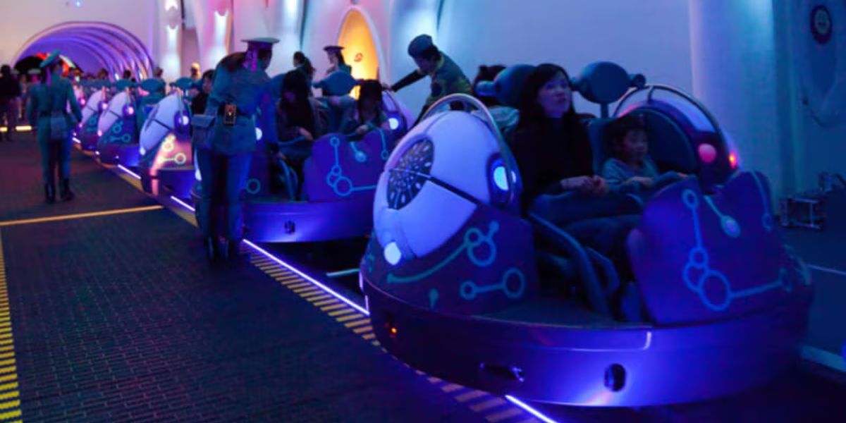 People are seated in futuristic, glowing rides in a dimly lit indoor amusement park setting at Universal Studios. The beloved ride resembles small pods on a track, and attendants in uniforms assist passengers. The atmosphere is vibrant with blue and purple lighting.