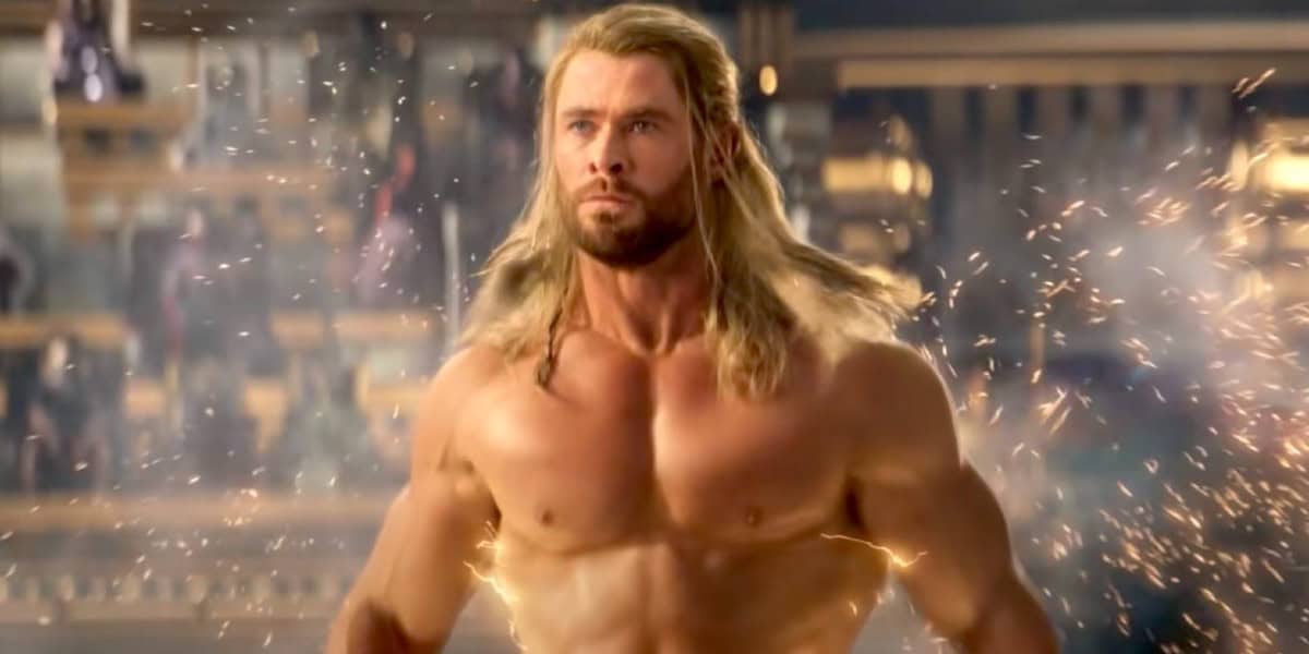 Thor (Chris Hemsworth) stands shirtless with sparks flying around him, set against a blurred background that suggests a mystical or ancient setting. His intense expression and the dramatic lighting highlight his powerful presence.