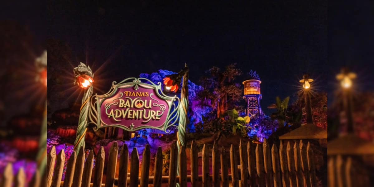 An amusement park attraction at night, illuminated in colorful lights. A large, ornate wooden sign reads "Tiana's Bayou Adventure," adorned with decorative elements. The background features elaborate rock formations and vegetation, with additional lighting.