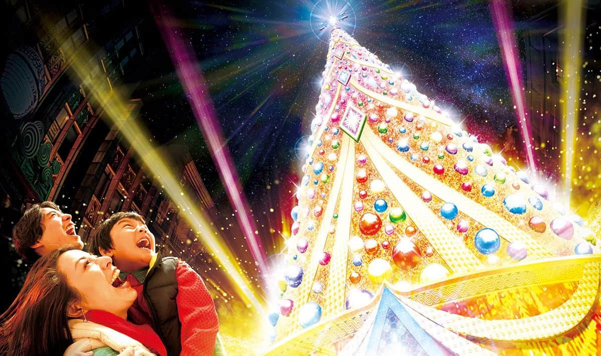 A group of people joyfully look up at a towering, brightly lit Christmas tree adorned with colorful ornaments, embodying the spirit of a universal Christmas celebration. The scene is vibrant with beams of light and a festive atmosphere, heralding the joy that 2024 promises to bring.