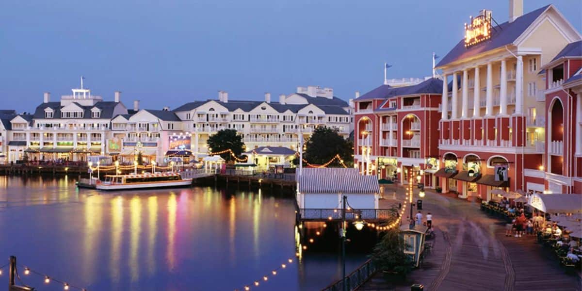 A scenic view of a waterfront boardwalk at dusk, featuring illuminated buildings with red and white exteriors reminiscent of a Cake Bake Shop, a calm body of water reflecting the lights, and a docked boat. Strings of lights adorn the walkway, creating an inviting atmosphere akin to Disney World.