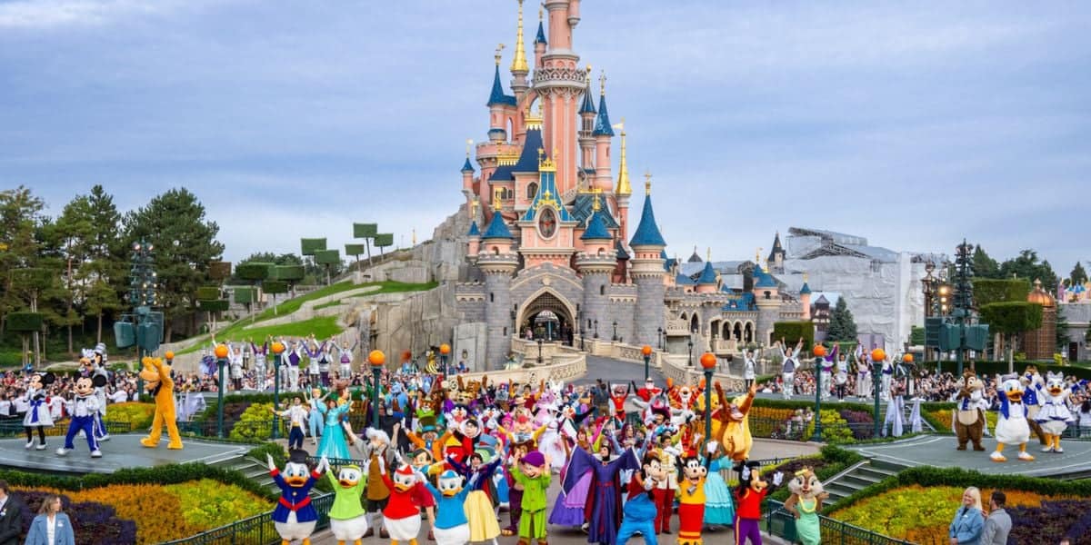 A vibrant parade at disneyland featuring numerous disney characters in colorful costumes, with a large, picturesque castle in the background and a crowd of attendees under a bright sky.