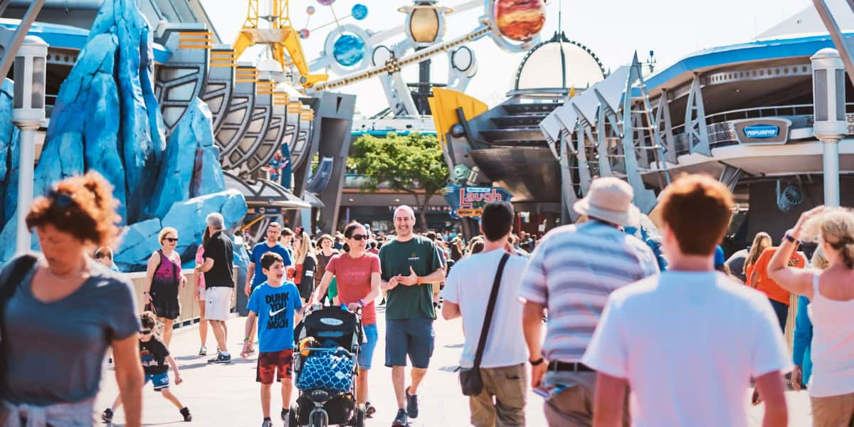 People walk through a busy, colorful theme park area with futuristic decor and attractions. Shops and rides line the path, and various families and individuals can be seen, some with strollers and others taking photos. The atmosphere is vibrant and lively.