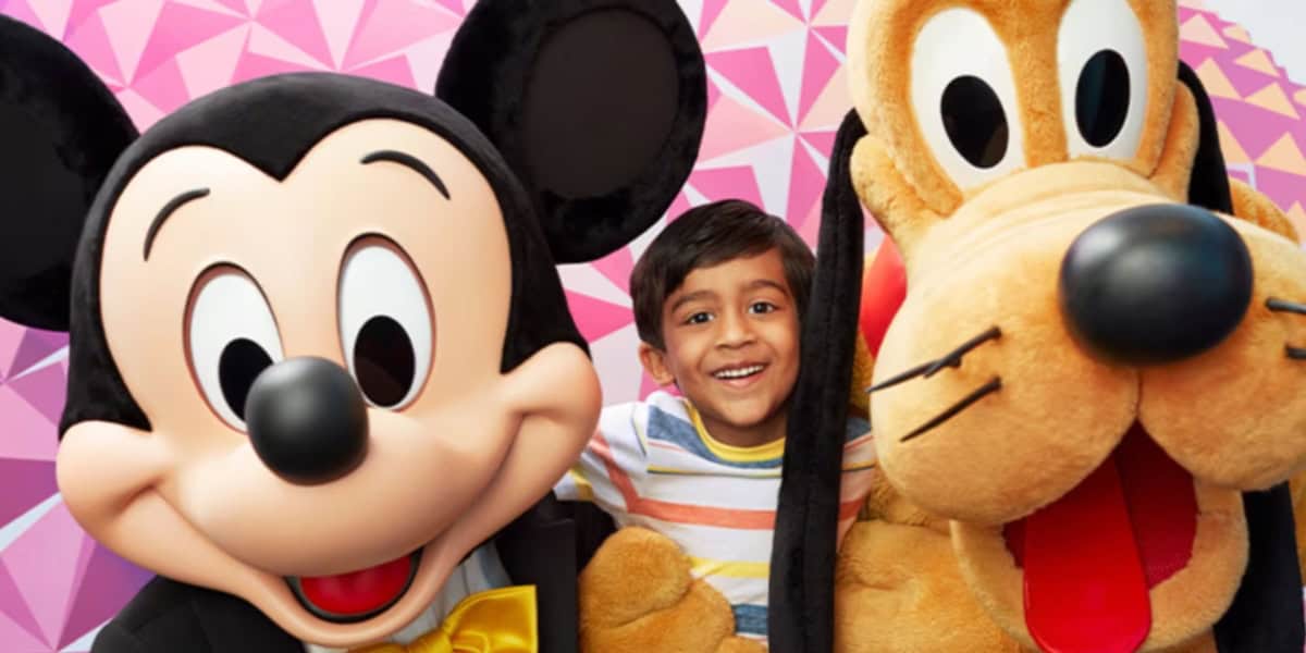 A young boy with a big smile is standing between two large, costumed characters—Mickey Mouse on the left and Pluto on the right. The background is a geometric pink pattern. They all appear happy and excited.