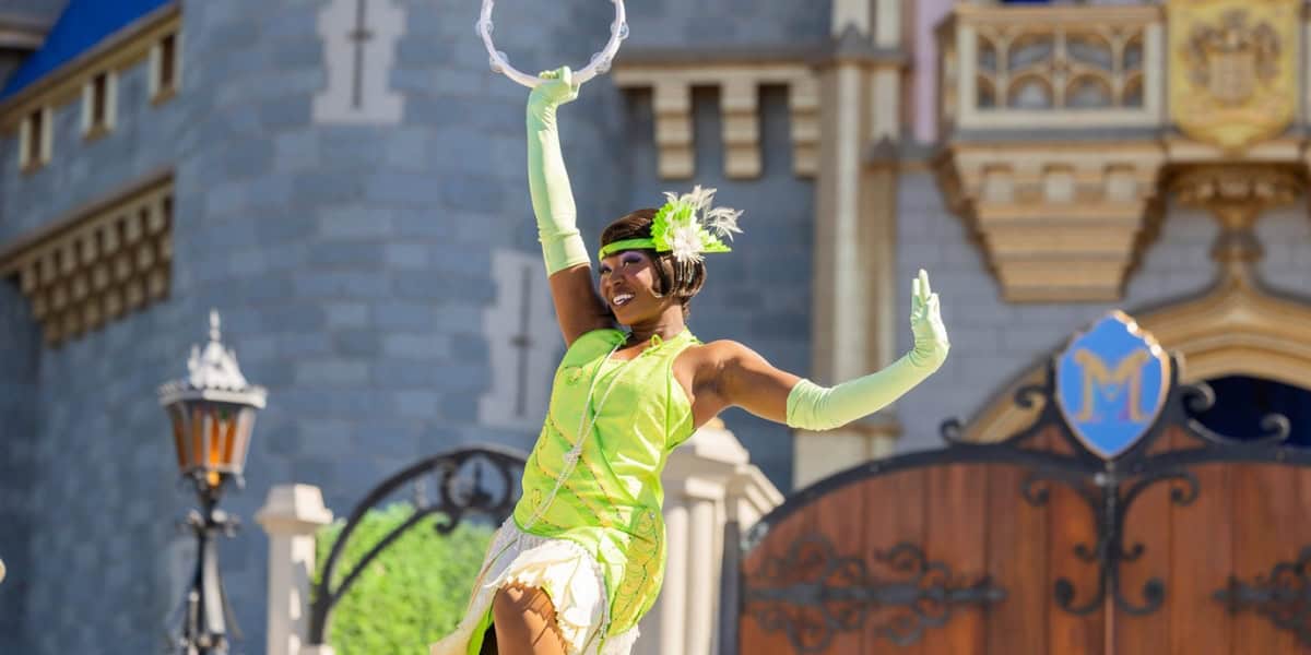 A performer dressed in a bright green costume with white gloves and a feathered headpiece holds a tambourine up high. They are standing in front of a castle with ornate details and a wooden door in the background.