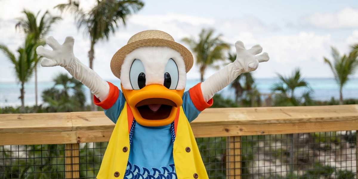 A person dressed in a Donald Duck costume stands on a wooden deck with arms wide open, wearing a yellow and blue outfit and a straw hat. Tropical palm trees and a clear blue sky with a body of water are visible in the background—your Disney Vacation Club adventure awaits!