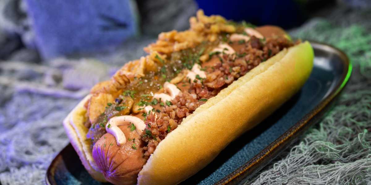 A close-up shot of a hot dog in a bun, topped with crisp fried onions, chopped bacon, a light drizzle of creamy sauce, and a sprinkling of herbs. Featured on the full list of food options at Mickey's Not-So-Scary Halloween Party 2024, it’s presented on a blue oval plate against a blurred background.