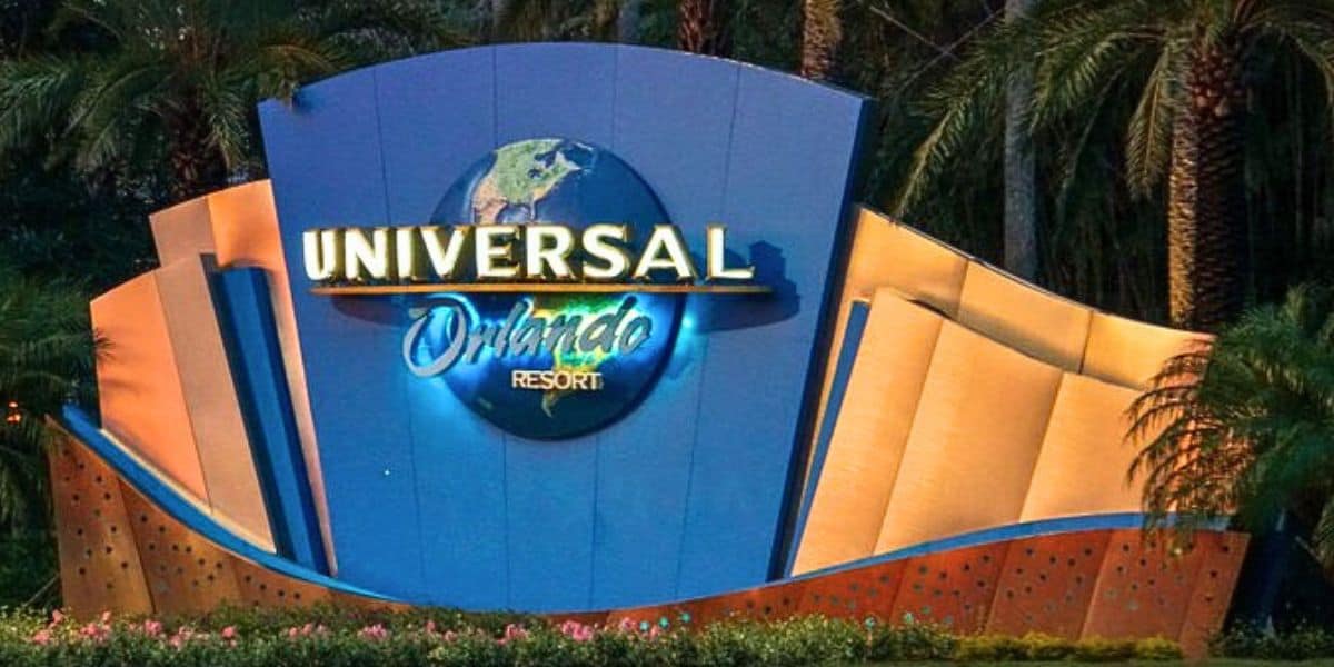 The image shows the entrance sign for Universal Orlando Resort. The large, colorful sign boasts a globe design and is surrounded by palm trees and greenery, creating a vibrant welcome that feels like an invitation to join a community celebration.