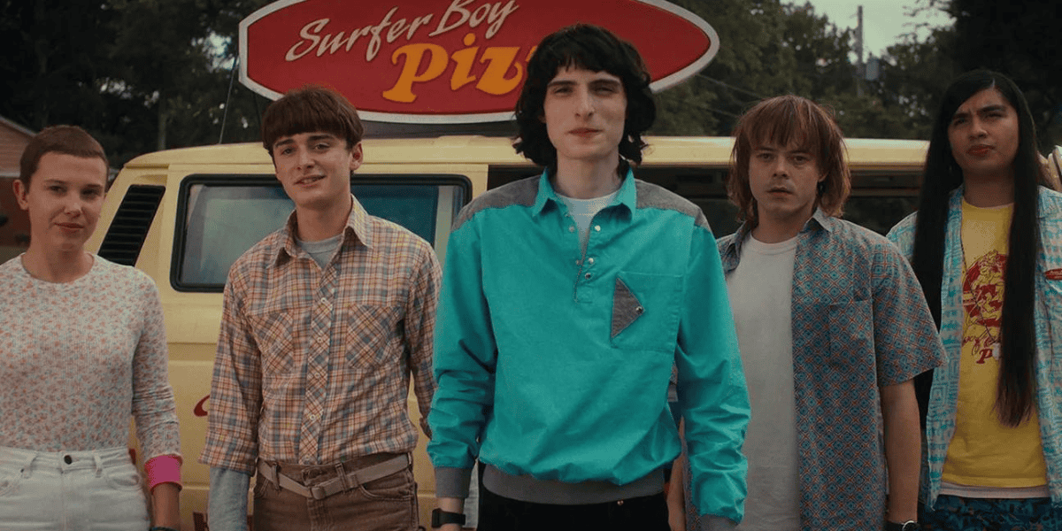 Five young individuals stand together in front of a yellow van with a "Surfer Boy Pizza" sign on top. They wear casual clothing, with a mix of retro styles, and appear to be looking ahead with neutral expressions. Trees and houses are visible in the background.