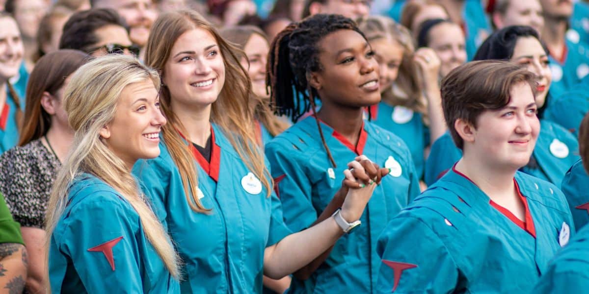 A diverse group of Disney cast members, mostly young adults, are standing closely together, smiling, and looking ahead. They are all wearing teal uniforms with badges. Some are holding hands, creating a sense of unity and celebration. The background shows more people in similar attire.