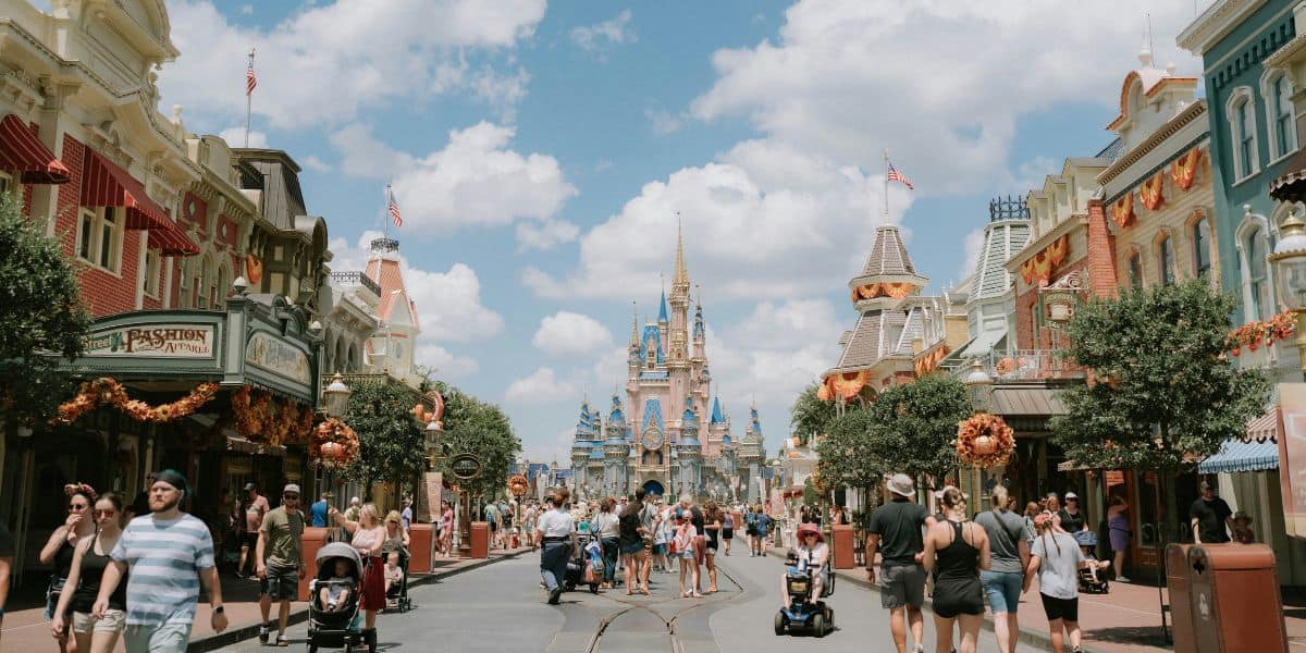 Main Street in the Disney theme park is bustling with visitors. People walk, push strollers, and some use mobility scooters. Decorated shops line the street, and a majestic castle stands in the background under a partly cloudy sky. Security ensures everyone enjoys their magical day safely.