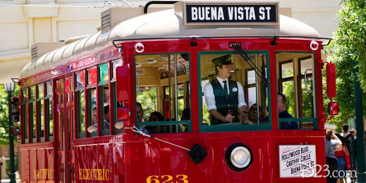 A red vintage trolley labeled "Buena Vista St" rolls through the tree-lined street at the Happiest Place on Earth. The uniformed driver, in a black hat and tie, is visible through the front window as passengers enjoy their ride inside. This charming scene captures Disney magic in motion.