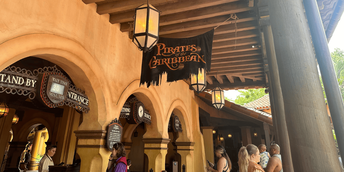 A group of people wait in line under hanging lanterns at the entrance to the Pirates of the Caribbean ride. The ride's sign, featuring tattered black cloth with orange text, is prominently displayed above the queue. The architecture is themed with arches and wooden beams.