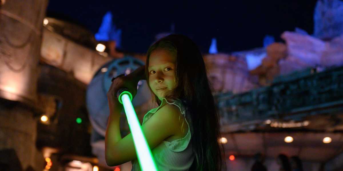 A young girl with long hair poses confidently holding a lit green lightsaber in an outdoor, nighttime setting at Walt Disney World’s Disney After Hours event. The background includes futuristic-looking structures and soft, colorful lighting, evoking a sci-fi or space-themed amusement park of 2025.