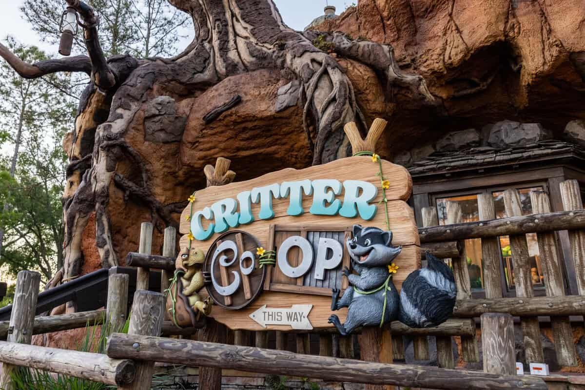 Entrance to "critter co-op" featuring whimsical decor with animal figures and rustic elements, surrounded by lush greenery and a large tree structure. signs point visitors in the direction to go inside of the Disney World Magic Kingdom.