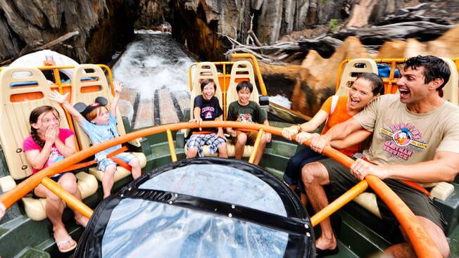 A group of six people on a circular raft ride are laughing and smiling as they navigate the rushing waters, inside of barge inside of Kali River Rapids at Disney World Animal Kingdom park. Surrounded by rocky terrain, everyone appears to be enjoying the thrilling experience.