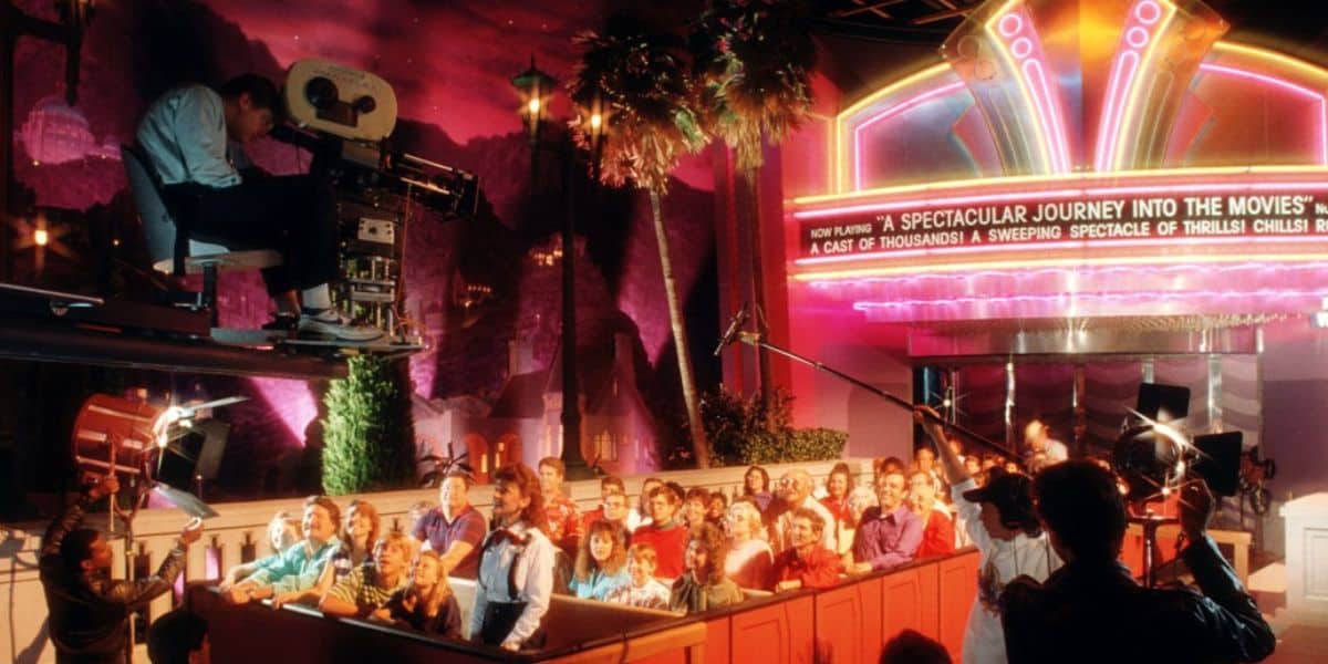 A crowd of people on a ride, surrounded by bright lights and neon signs, simulating a movie set experience. A Disney Animatronic camera operator is elevated on the left, capturing the scene. The atmosphere is vibrant, with a mix of palm trees and pink lighting in the background.