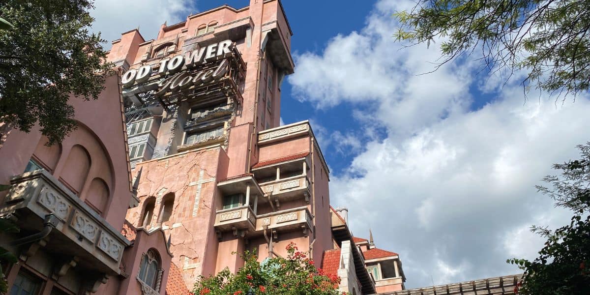 A tall, aged, pinkish-brown building with a sign reading "Hollywood Tower Hotel" stands against a bright blue sky with white clouds. With its old-fashioned design and worn-out facades surrounded by greenery, one might imagine Disney World characters exploring its partially exposed brickwork.