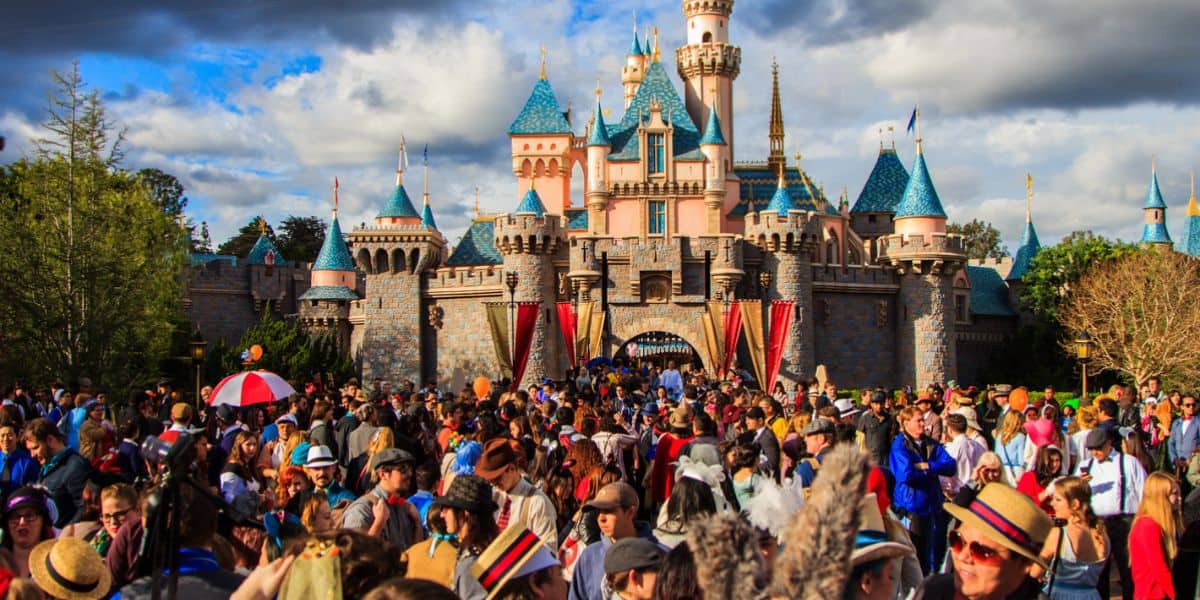 A large crowd is gathered in front of a colorful castle with blue turrets under a partly cloudy sky. Many people are wearing hats and themed clothes, enjoying a lively and vibrant atmosphere. Trees and landscape surround the scene.