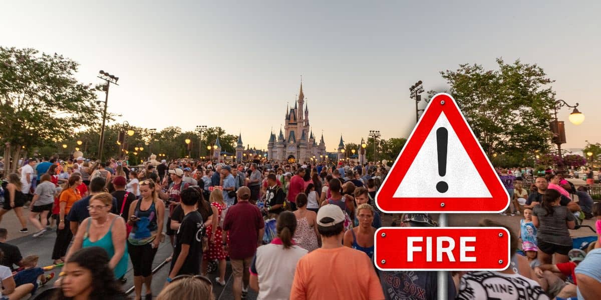 A large crowd is gathered in front of a castle, with a prominent warning sign in the foreground. The sign displays a red triangle with an exclamation mark and a red rectangle that reads "FIRE." The scene is outdoors at dusk at Walt Disney World.