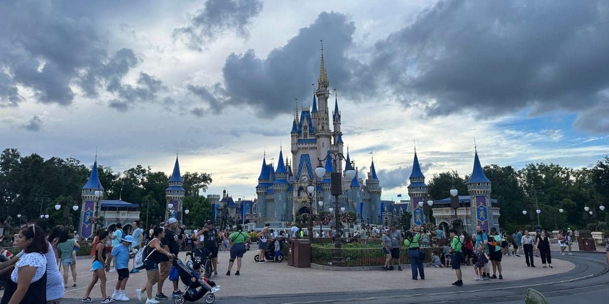 Crowds of people walk in front of a grand castle with blue accents and spires, framed by lush greenery under a cloudy sky. The lively, colorful scene evokes a Disney World atmosphere.