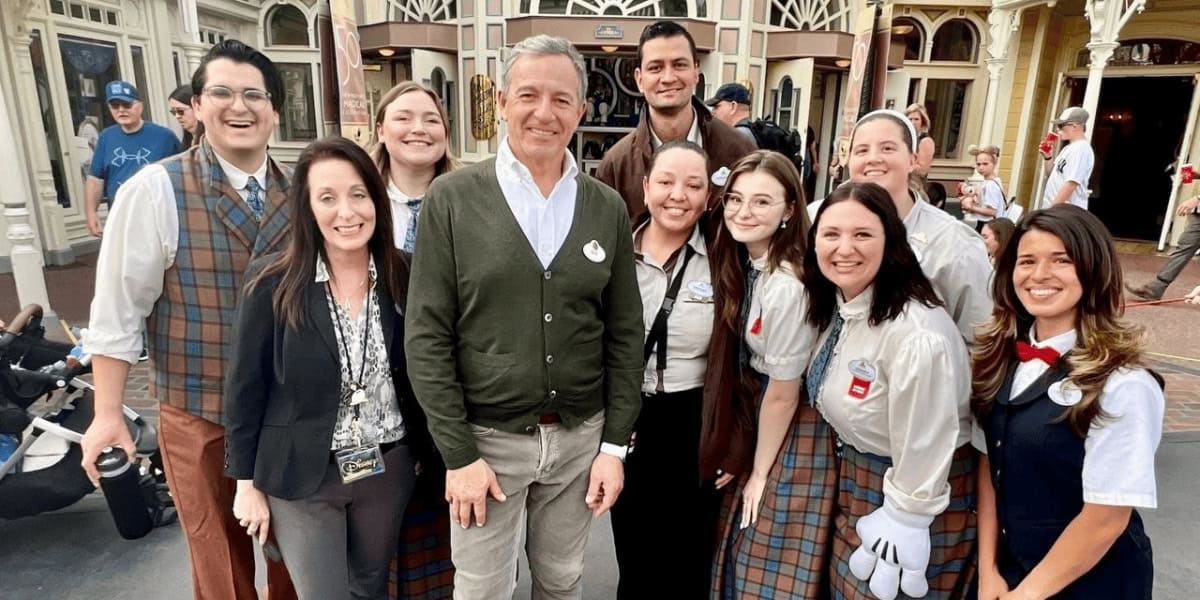 A group of people smiling and posing together in an outdoor setting, with a mix of casual and themed clothing. The background shows a building with decorative elements reminiscent of Bob Iger's visionary style. The atmosphere is cheerful and friendly.