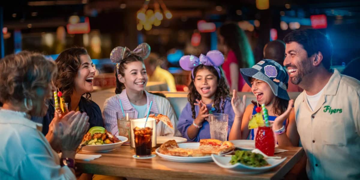 A family of six sits around a restaurant table, laughing and enjoying a meal. The table is set with various dishes, including fries, pizza, and drinks. Two young girls wear colorful mouse ear headbands, adding to the festive atmosphere.