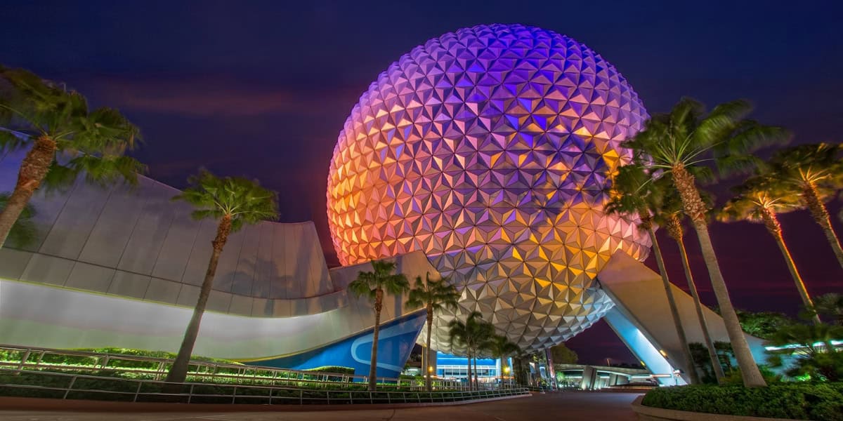 Night view of Epcot's iconic Spaceship Earth geodesic sphere at Walt Disney World, Florida, illuminated with colorful lights in shades of purple, blue, and orange. Palm trees line the entrance as Disney World rides buzz nearby, and the sky is deep dusk, adding to the vibrant atmosphere.