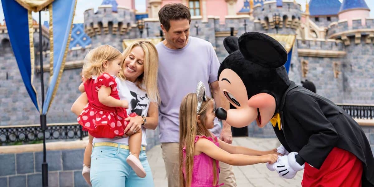 A family of four, including two young children, interacts with Mickey Mouse in front of Sleeping Beauty Castle at Disneyland.